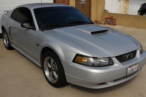 2002 Ford Mustang - A silver 2002 Ford Mustang GT coupe is parked on a concrete driveway in front of a brick building. The front passenger side of the car is a…