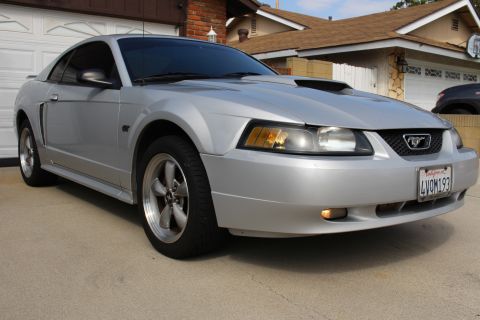 2002 Ford Mustang - A silver Ford Mustang GT is parked on a concrete driveway in front of a residential garage. The car is viewed from a low, three-quarter fro…
