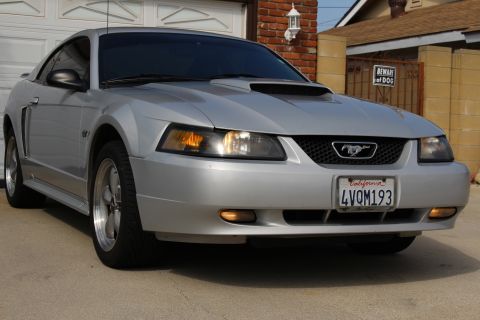 2002 Ford Mustang - The front driver's side quarter view of a silver Ford Mustang is parked on a concrete driveway in front of a garage and house. The car has…