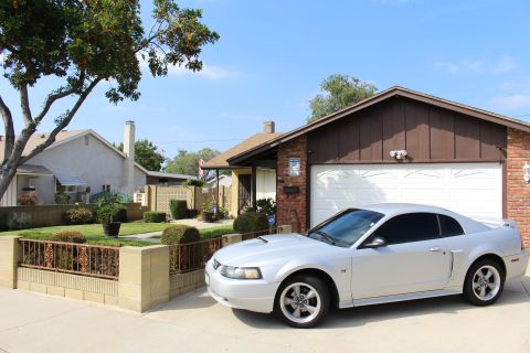 2002 Ford Mustang - A silver 2002 Ford Mustang GT coupe is parked in the driveway of a brick and wood-paneled house with a white garage door. The car is viewed…