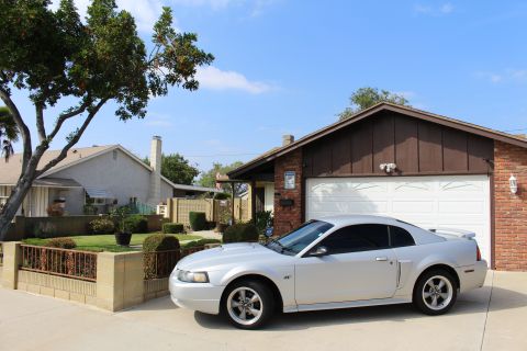 2002 Ford Mustang - A silver Ford Mustang coupe is parked in front of a brick and wood-sided garage on a sunny day. The car is viewed from the front three-quar…
