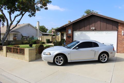 2002 Ford Mustang - A silver Ford Mustang coupe is parked in the driveway of a suburban house. The car is viewed from the front 3/4 angle, showing its side and…