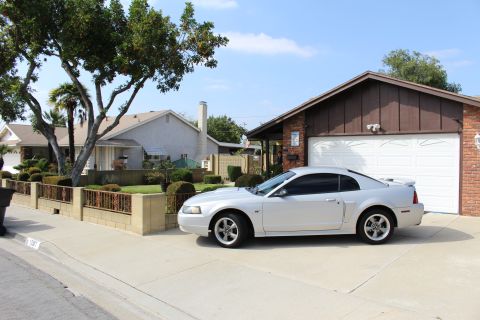2002 Ford Mustang - A silver Ford Mustang coupe is parked in the driveway of a suburban home. The car is viewed from the side, with a white garage door and bri…