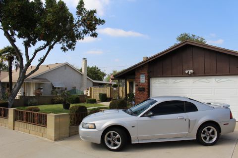 2002 Ford Mustang - A silver Ford Mustang coupe is parked in a driveway in front of a brick and wood-sided house with a white garage door. The car is viewed fr…