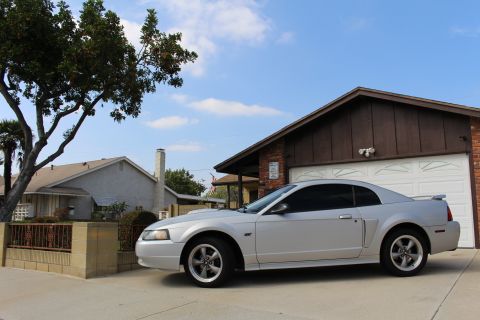 2002 Ford Mustang - A silver Ford Mustang coupe is parked in a driveway in front of a brick and wood-paneled garage. The car is viewed from the side, with a la…