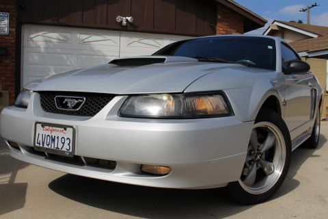 2002 Ford Mustang - The front passenger side quarter view of a silver 2002 Ford Mustang is parked in front of a garage. The car features a hood scoop, a black…