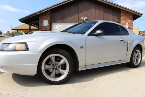 2002 Ford Mustang - The side view of a silver Ford Mustang GT coupe is parked in front of a brick and wood-paneled garage. The car features tinted windows and…