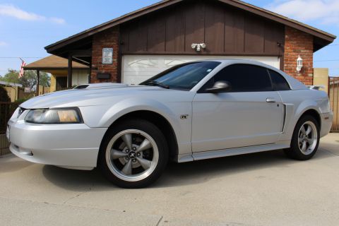 2002 Ford Mustang - A silver Ford Mustang GT coupe is parked in front of a brick and wood-paneled garage. The car is viewed from the front three-quarter angle,…