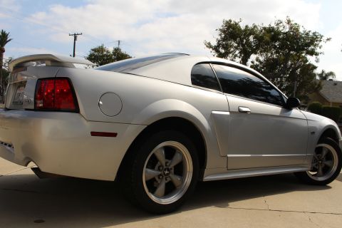 2002 Ford Mustang - The rear passenger side of a silver Ford Mustang is shown from a low angle, parked on a concrete surface with trees and a cloudy sky in the…