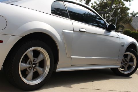 2002 Ford Mustang - The side profile of a silver Ford Mustang is shown, focusing on the rear wheel and door. The car features tinted windows and a GT emblem on…