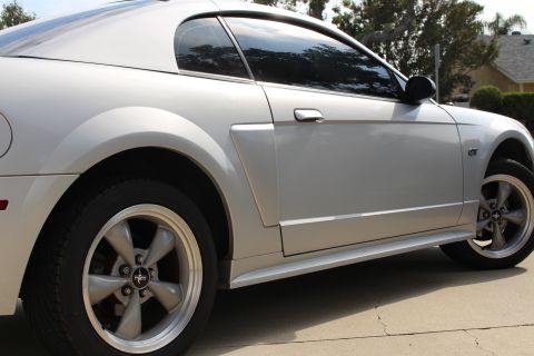 2002 Ford Mustang - The side profile of a silver Ford Mustang coupe is shown, parked on a concrete surface with trees and a house in the background.