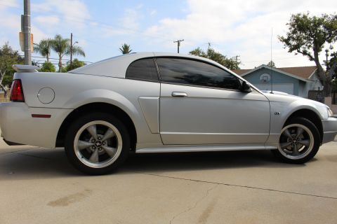 2002 Ford Mustang - The side view of a silver Ford Mustang coupe is parked on a concrete driveway. The car has tinted windows, a rear spoiler, and five-spoke a…