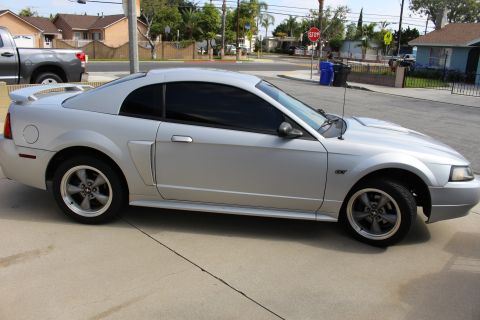 2002 Ford Mustang - The side view of a silver 2002 Ford Mustang GT coupe is parked on a concrete driveway in front of a residential area. The car has tinted wi…