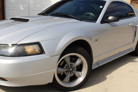 2002 Ford Mustang - The front quarter panel and wheel of a silver Ford Mustang GT are shown. The car features a hood scoop and a GT badge on the fender.