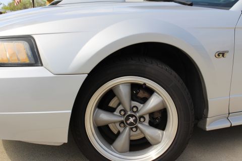 2002 Ford Mustang - Close-up of the front passenger side wheel and fender of a silver Ford Mustang. The image shows a silver five-spoke wheel with a black and…