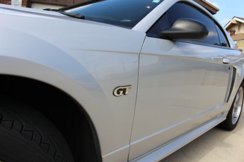 2002 Ford Mustang - A close-up, side view of a silver Ford Mustang GT's front fender and door. The distinctive GT badge is clearly visible on the fender.