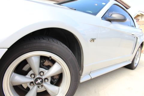 2002 Ford Mustang - A close-up, low-angle shot of the front passenger side of a white Ford Mustang GT. The image focuses on the wheel, tire, and fender, with t…
