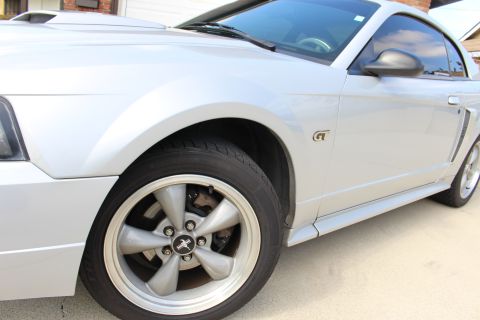 2002 Ford Mustang - A close-up, low-angle shot of the front passenger side wheel and fender of a silver Ford Mustang GT. The car is parked on a concrete drivew…