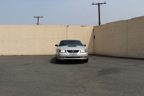 2002 Ford Mustang - Front view of a silver Ford Mustang parked in front of a tan brick wall. The vehicle has its headlights on and a license plate reading "4VO…