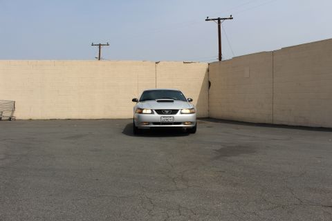 2002 Ford Mustang - A silver Ford Mustang is parked in front of a tan brick wall under a clear sky. The car is facing the camera, and a shopping cart is visibl…