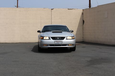 2002 Ford Mustang - Front view of a silver Ford Mustang parked in front of a tan brick wall. The car has its headlights on and displays a California license pl…