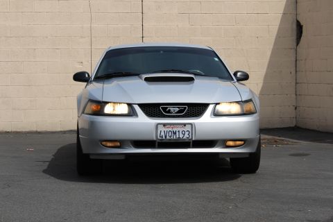 2002 Ford Mustang - Front view of a silver Ford Mustang parked in front of a light-colored brick wall. The car has its headlights on and displays a California…