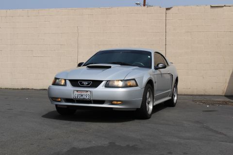 2002 Ford Mustang - A silver 2002 Ford Mustang GT is parked in front of a beige brick wall. The front of the car faces the camera, showing its headlights, gril…