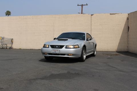 2002 Ford Mustang - A silver Ford Mustang is parked in front of a tan brick wall. The car is facing the camera at a slight angle, showing its front grille, hea…
