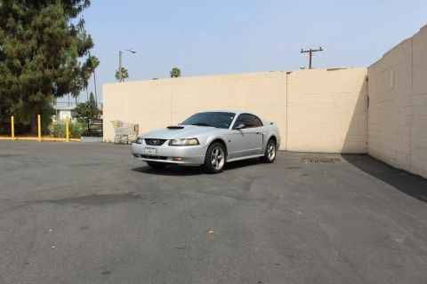 2002 Ford Mustang - A silver Ford Mustang coupe is parked in an asphalt lot next to a light-colored brick wall. The car is viewed from a 3/4 front angle, showi…