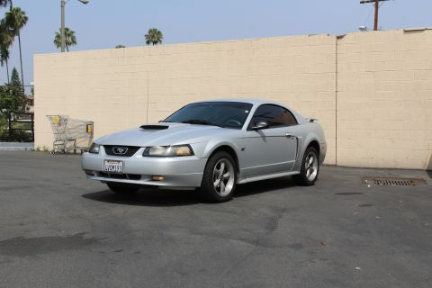 2002 Ford Mustang - A silver 2002 Ford Mustang coupe is parked in front of a beige brick wall. A shopping cart is visible to the left of the vehicle.