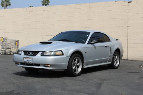 2002 Ford Mustang - A silver 2002 Ford Mustang GT is parked in front of a tan brick wall. The car is viewed from a three-quarter front angle, showing its front…