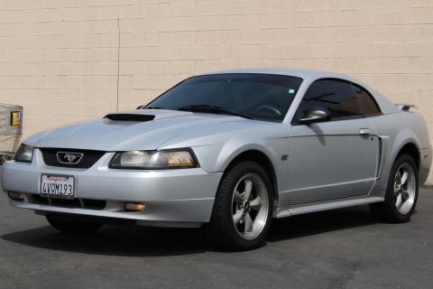 2002 Ford Mustang - A silver 2002 Ford Mustang GT coupe is parked outdoors in front of a brick wall. The car is viewed from a 3/4 front angle, showing its hood…