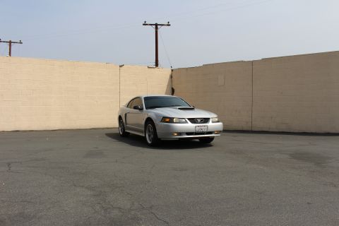 2002 Ford Mustang - A silver Ford Mustang coupe is parked in an asphalt lot in front of a light-colored brick wall. The car is facing slightly to the right, sh…