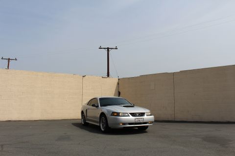 2002 Ford Mustang - A silver Ford Mustang coupe is parked in front of a light-colored brick wall under a hazy sky. The car is facing the camera at a slight ang…