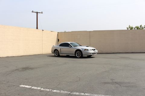 2002 Ford Mustang - A silver Ford Mustang coupe is parked in an asphalt lot in front of a beige brick wall. A wooden utility pole stands to the left of the veh…