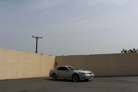 2002 Ford Mustang - A silver 2002 Ford Mustang coupe is parked in an asphalt lot next to a tan brick wall under a hazy sky. A wooden utility pole with wires st…