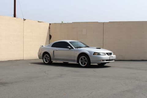 2002 Ford Mustang - A silver 2002 Ford Mustang GT coupe is parked on asphalt in front of a tan brick wall. The car is viewed from the side at a 3/4 front angle.