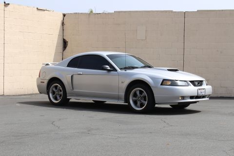 2002 Ford Mustang - A silver 2002 Ford Mustang GT coupe is parked on asphalt in front of a beige brick wall. The car is viewed from a 3/4 front angle, showing…