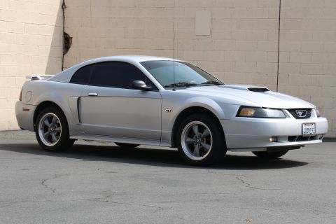 2002 Ford Mustang - A silver 2002 Ford Mustang GT coupe is parked on asphalt in front of a light-colored brick wall. The car has tinted windows and a rear spoi…