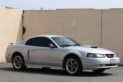 2002 Ford Mustang - A silver 2002 Ford Mustang GT coupe is parked in front of a textured, light-colored brick wall. The car is viewed from the front three-quar…