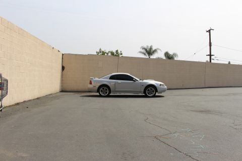 2002 Ford Mustang - A silver 2002 Ford Mustang coupe is parked in an asphalt lot next to a beige brick wall. The car is viewed from the side, with a shopping c…