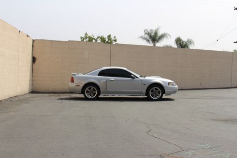2002 Ford Mustang - A silver 2002 Ford Mustang coupe is parked in front of a light-colored brick wall. The car is viewed from the side, showing its profile and…