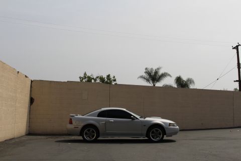 2002 Ford Mustang - A silver 2002 Ford Mustang coupe is parked in front of a tan brick wall under a hazy sky. The car is viewed from the side, showing its prof…