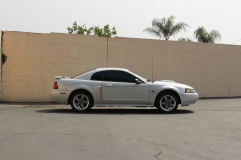 2002 Ford Mustang - Side profile of a silver 2002 Ford Mustang coupe parked on asphalt in front of a tan brick wall. The car features tinted windows and a rear…