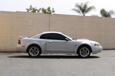 2002 Ford Mustang - Side profile of a silver Ford Mustang coupe parked in front of a textured tan brick wall with palm trees visible above. The car has tinted…