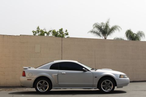 2002 Ford Mustang - Side view of a silver Ford Mustang coupe parked in front of a tan brick wall. The car has tinted windows and a spoiler on the trunk.