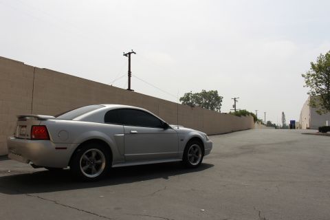 2002 Ford Mustang - A silver 2002 Ford Mustang coupe is parked on an asphalt lot next to a brick wall.
