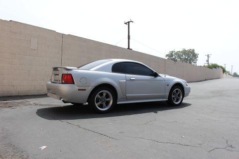 2002 Ford Mustang - A silver 2002 Ford Mustang coupe is parked on an asphalt lot next to a beige wall.
