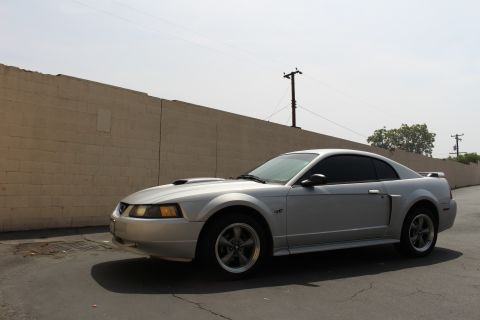 2002 Ford Mustang - A silver 2002 Ford Mustang GT is parked on a street next to a tan brick wall under a bright sky.