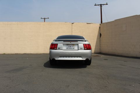 2002 Ford Mustang - This silver Ford Mustang is parked in an outdoor lot with a tan brick wall behind it. The car has a spoiler and dual exhaust pipes.